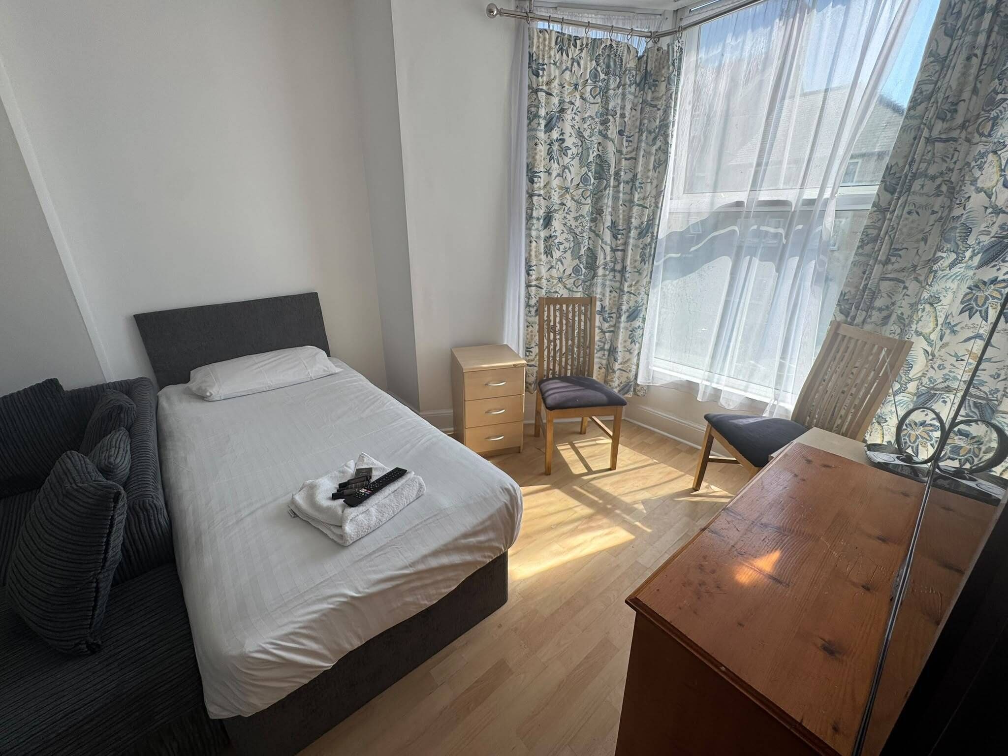 A bedroom with a bed, a wooden nightstand, a chair, a desk, and a window with curtains.