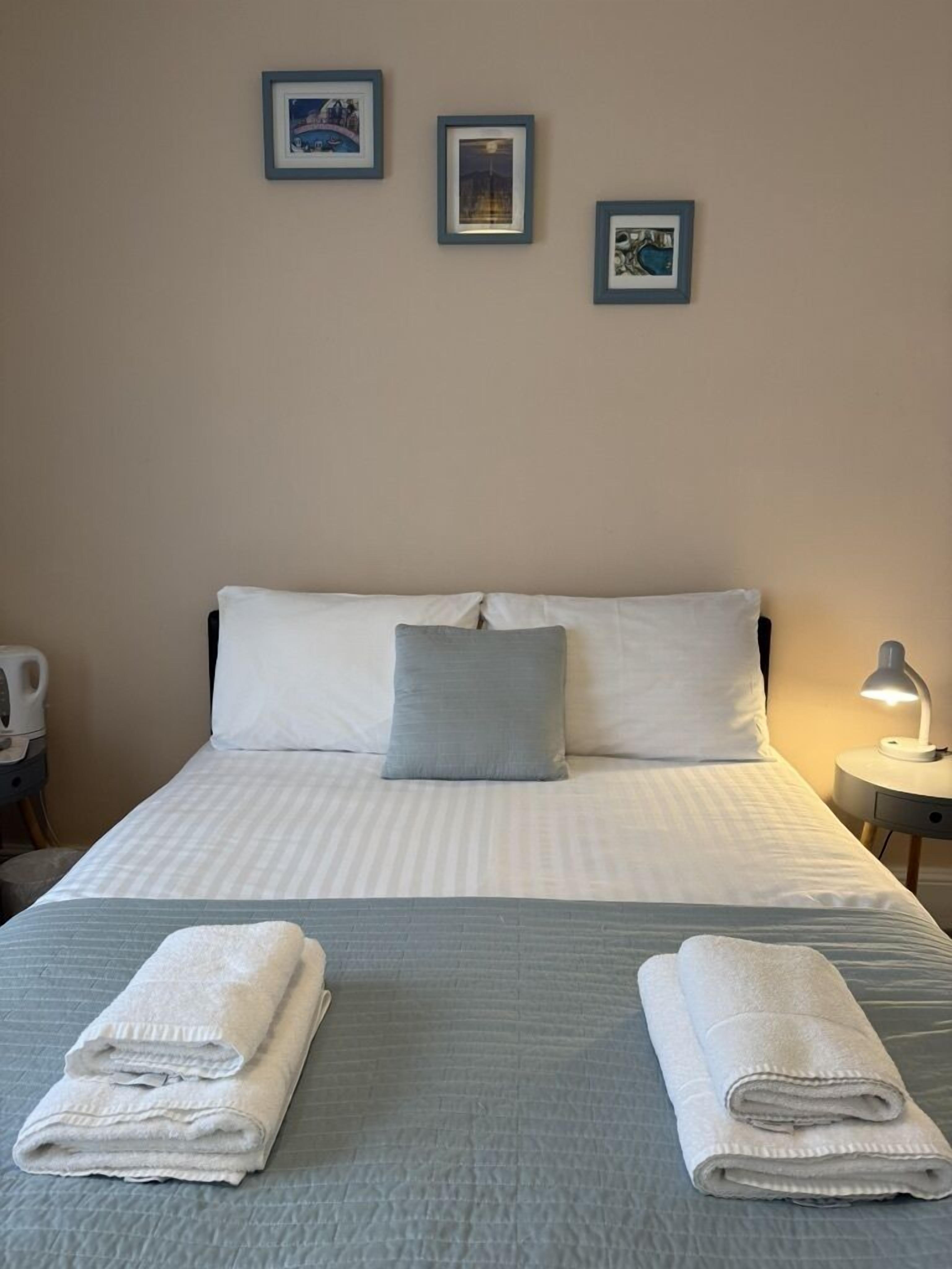 A neatly made bed with white and gray bedding, two pillows, and two folded towels on the bed. There are three framed pictures on the wall above the bed.
