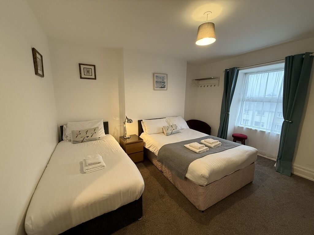 A hotel room with two single beds, a small table, a nightstand, a lamp, and a window with curtains.