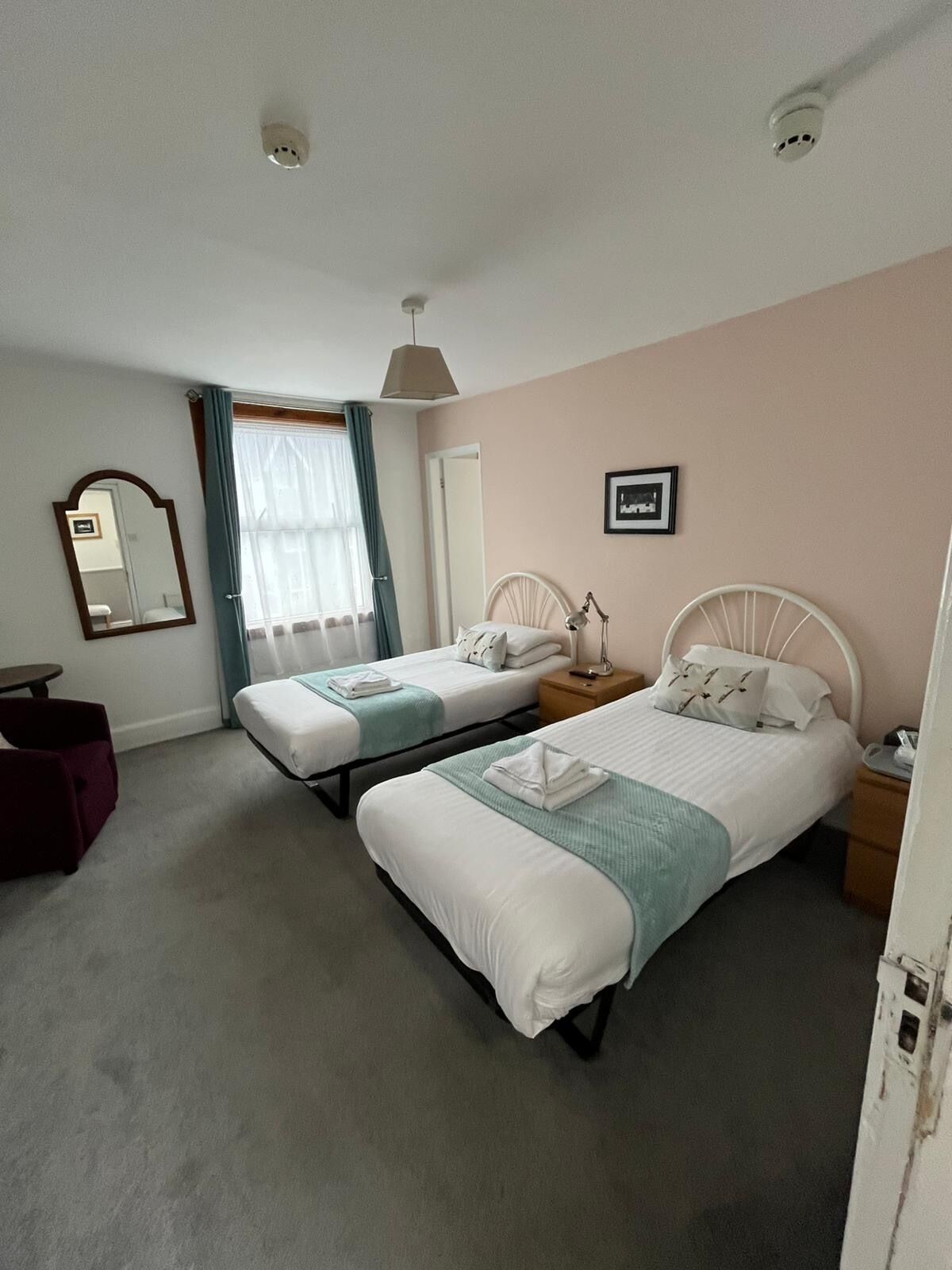 Une chambre d’hôtel avec deux lits simples, une petite table, une chaise, un miroir et une fenêtre avec des rideaux.