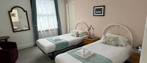 A hotel room with two single beds, a small table, a chair, a mirror, and a window with curtains.