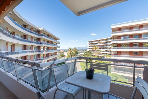 Un balcon avec une table et des chaises, donnant sur un immeuble à plusieurs étages, lui aussi doté de balcons, et sur un ciel dégagé.