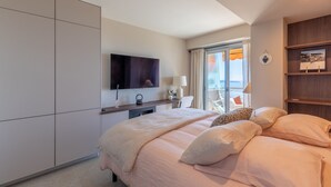Ein modernes Hotelzimmer mit einem großen Bett, einem Flachbildfernseher, einem Schreibtisch mit Lampe und Blick ins Freie durch große Fenster.