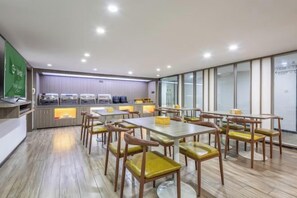 A modern dining area with multiple tables and chairs, a buffet counter, and a large window providing natural light. - Hilbert Hotel (Guangzhou Nansha Wanda Plaza Jinzhou Subway Station) (Guangzhou)