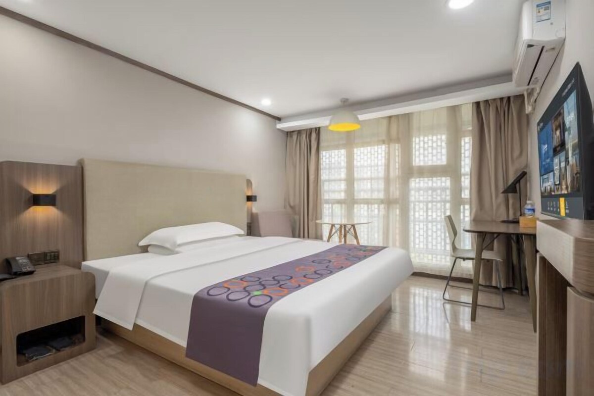Quarto de hotel com cama, mesa de cabeceira, luminária, escrivaninha, cadeira e janela com cortinas.