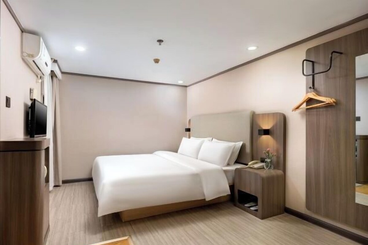 A modern hotel room with a large bed, a bedside table, a TV, and a wooden wardrobe.