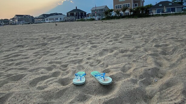 Flip-flops på en sandstrand med huse i baggrunden.