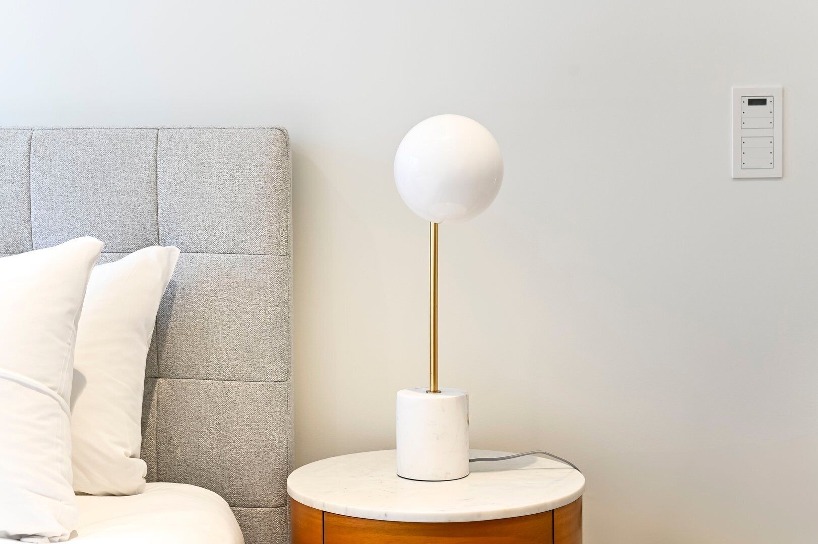 A bed with white bedding, a grey upholstered headboard, and a bedside table with a white lamp and a power outlet.