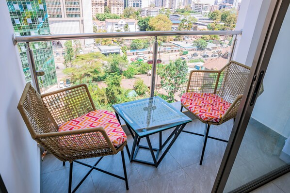 Een balkon met twee rieten stoelen en een kleine tafel, met uitzicht op een stadsgezicht met gebouwen en groen.