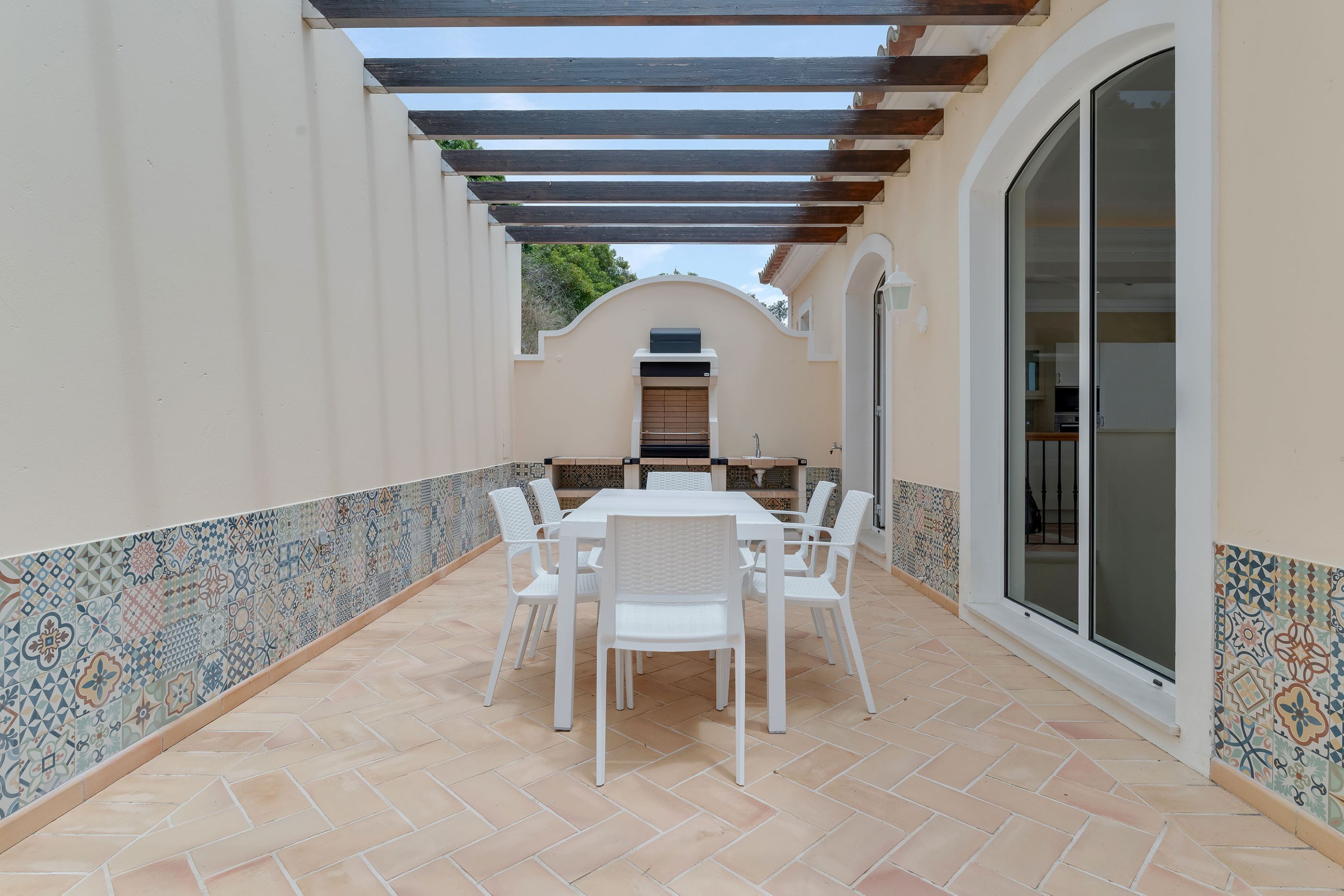 Une terrasse couverte avec un sol carrelé, une table blanche avec des chaises et une vue sur l’intérieur à travers une porte vitrée.