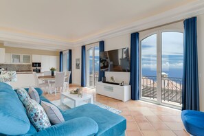Un salon moderne avec un canapé bleu, une table basse blanche et un téléviseur. Il y a un balcon avec vue sur la mer.