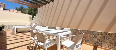 Een terras buiten met witte tafels en stoelen, een ingebouwde barbecue en een betegelde muur met een geometrisch patroon.