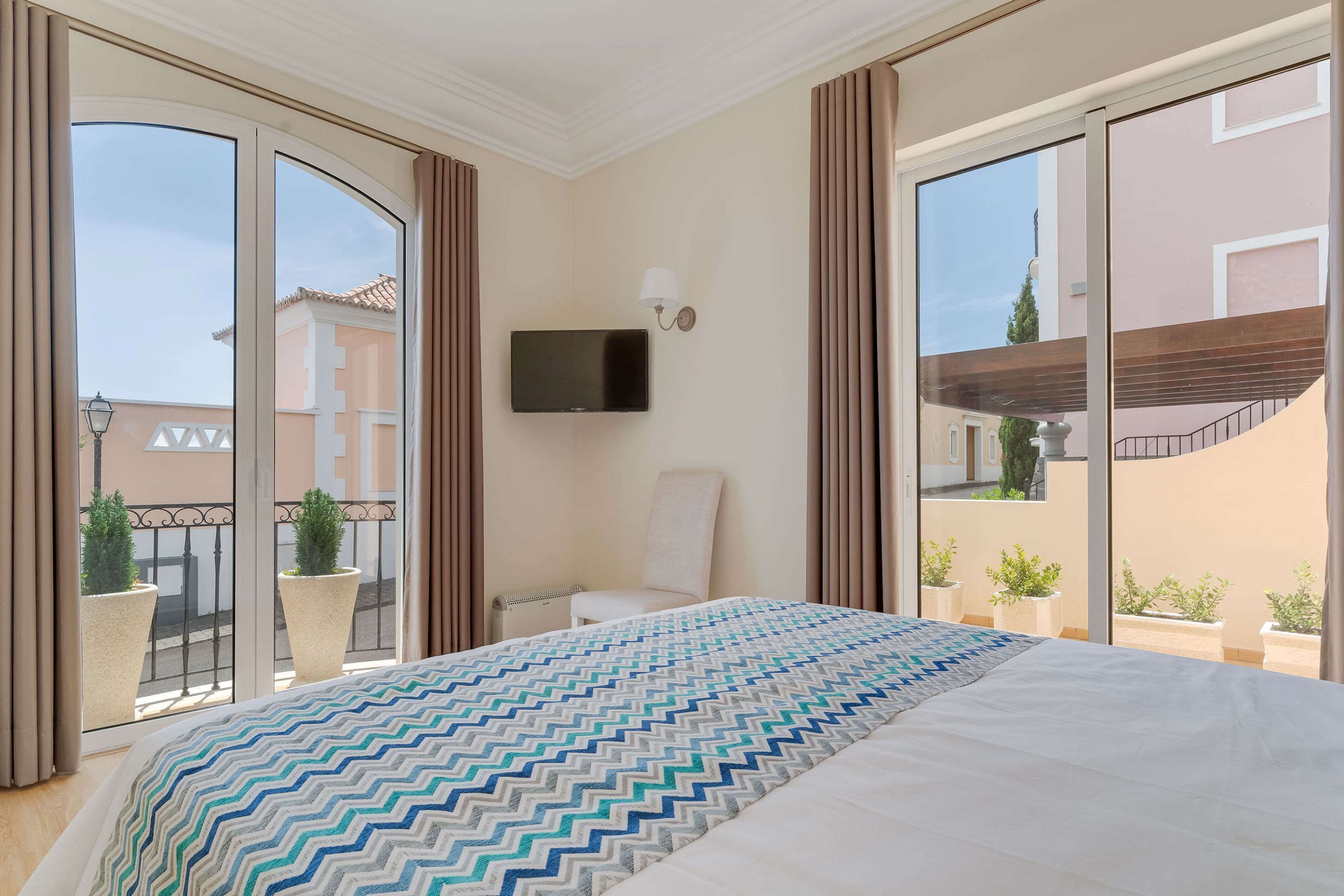 A hotel room with a large bed, a television mounted on the wall, and a balcony with potted plants.