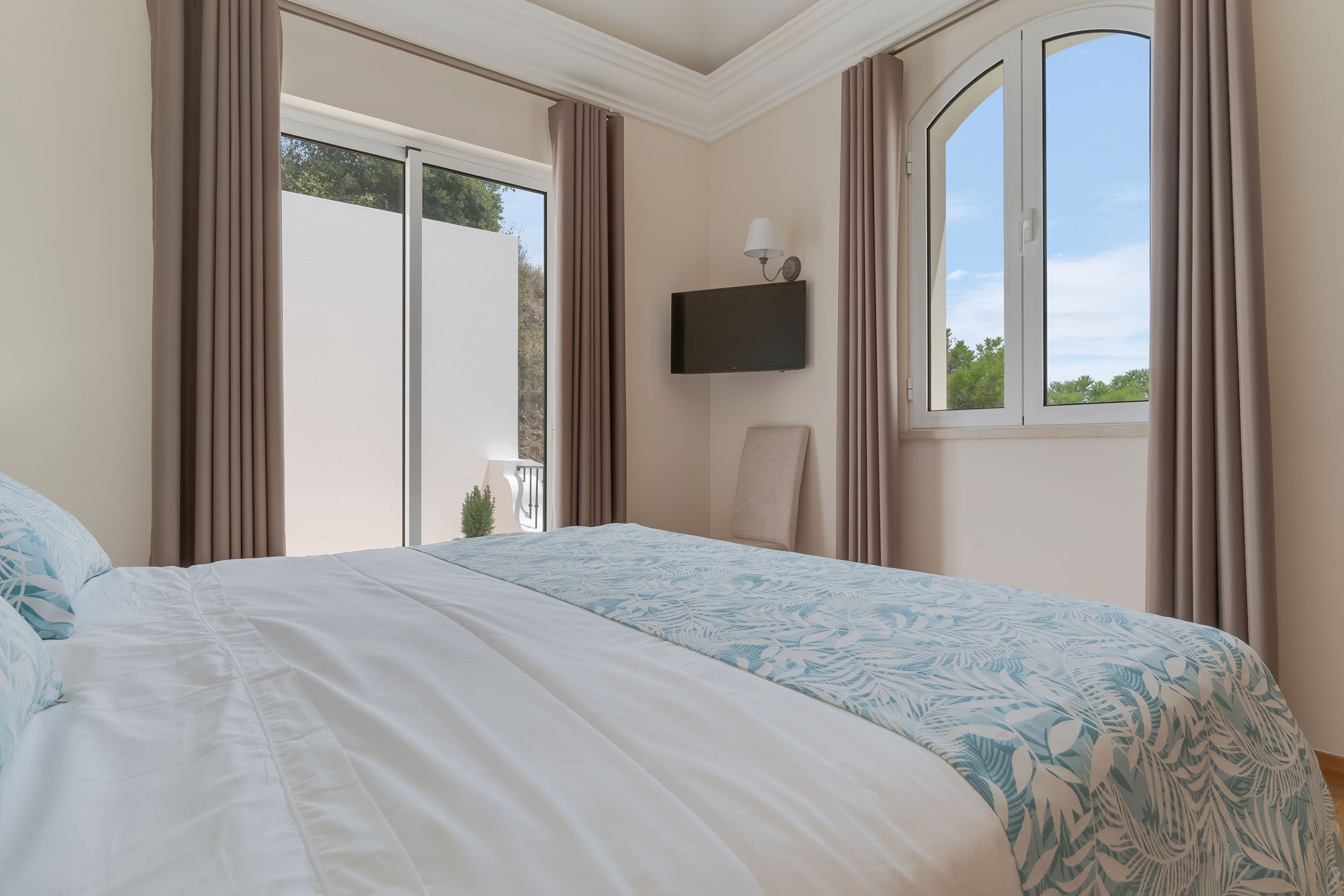 Un dormitorio con una cama grande, un televisor montado en la pared y un ventanal con cortinas.