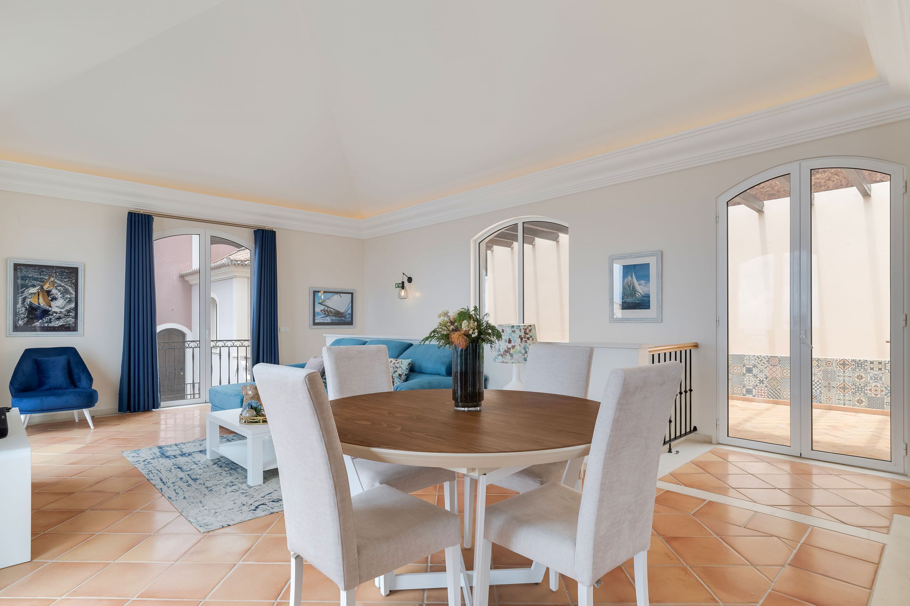 Ein modernes Wohnzimmer mit einem runden Holztisch, beigen Sesseln, einem blauen Sofa und einem blauen Sessel. Ein großes Fenster mit Vorhängen, ein Gemälde an der Wand und ein Fliesenboden sind ebenfalls zu sehen.