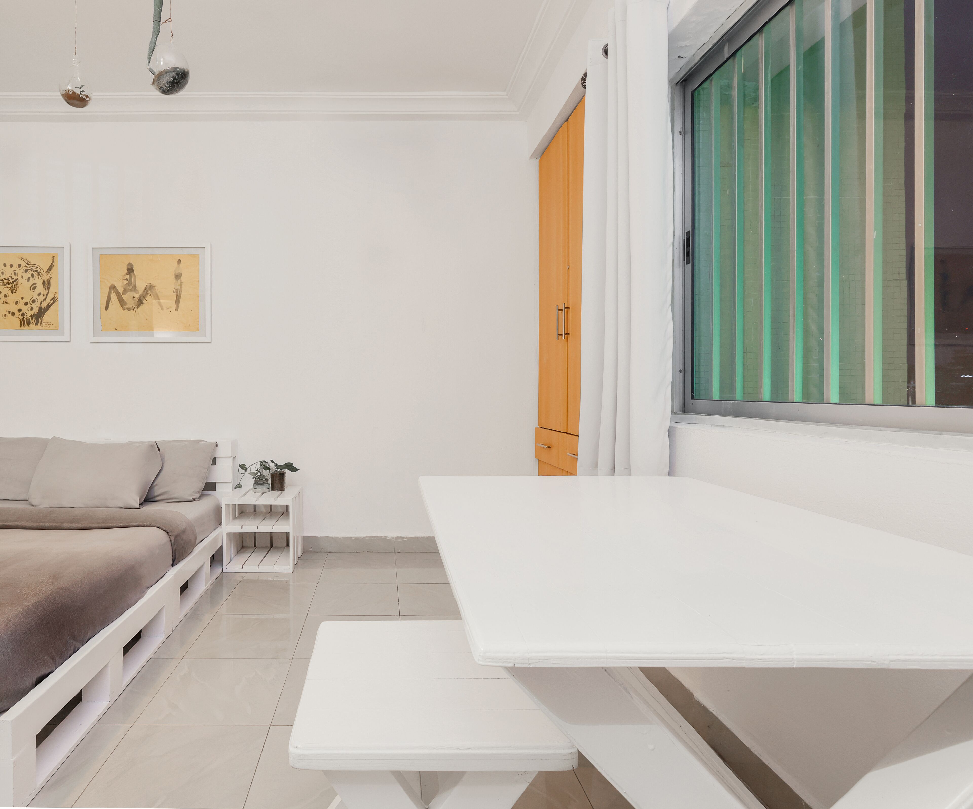 A minimalist room with a white bed, a white table, and a window with green vertical blinds.