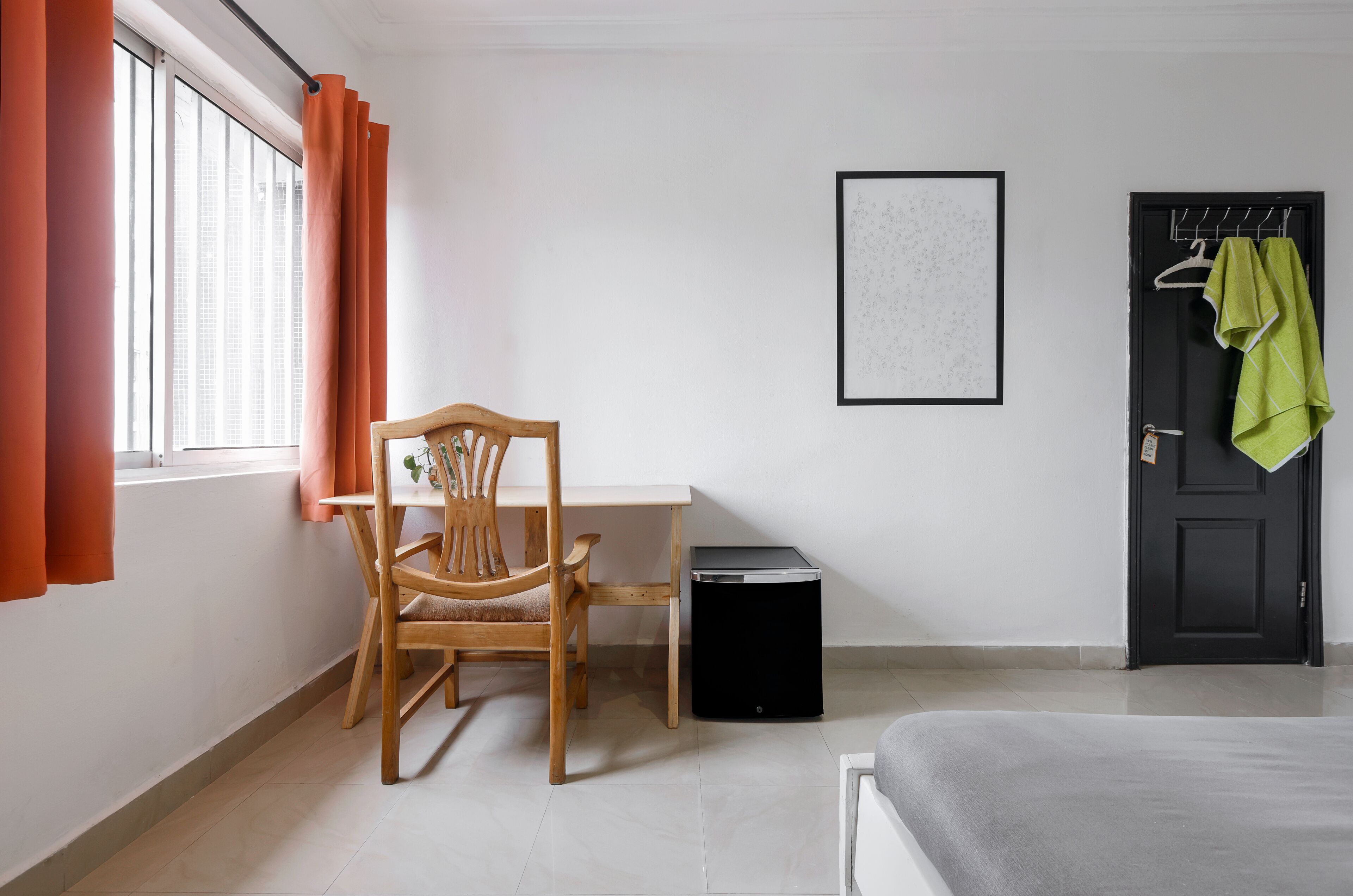 Ein kleines Hotelzimmer mit Holzstuhl, Tisch, schwarzem Papierkorb, einem gerahmten Bild an der Wand und einer Tür, an deren Griff ein Handtuch hängt.