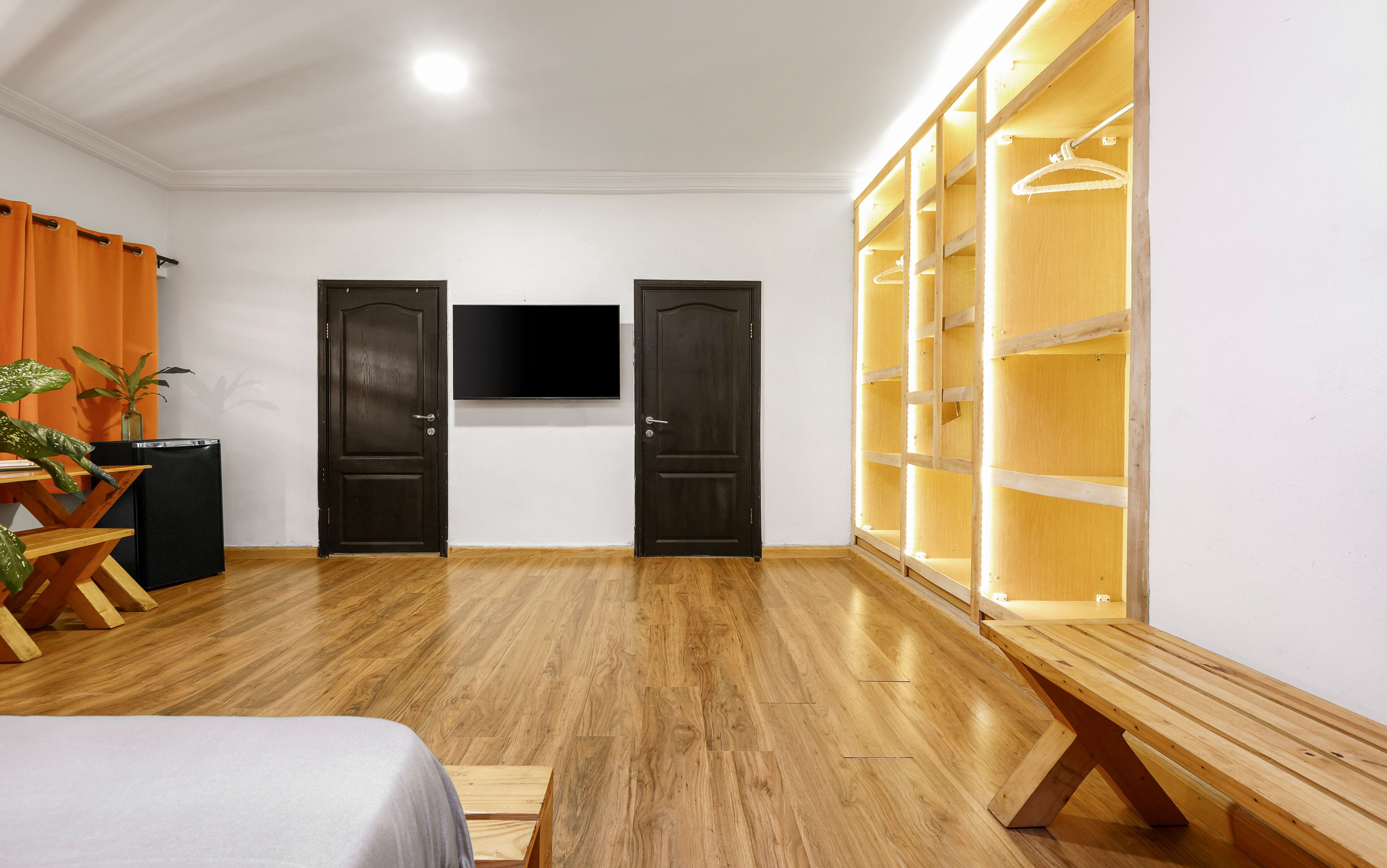 Quarto com piso de madeira, duas portas de madeira escura, uma TV de tela plana na parede e um guarda-roupa de madeira com espaço para pendurar roupas.