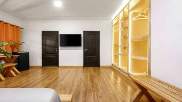 Quarto com piso de madeira, duas portas de madeira escura, uma TV de tela plana na parede e um guarda-roupa de madeira com espaço para pendurar roupas.