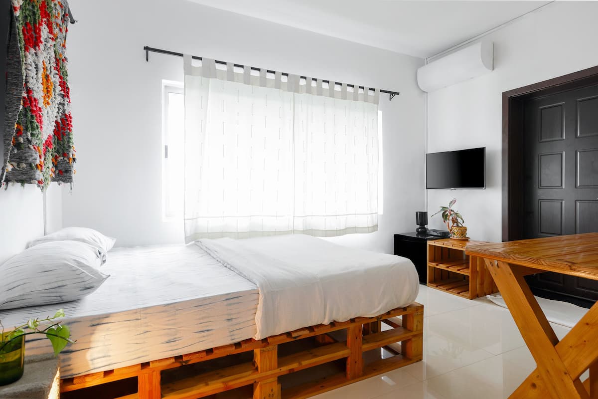 A bedroom with a wooden bed, a TV mounted on the wall, and a window with sheer curtains.
