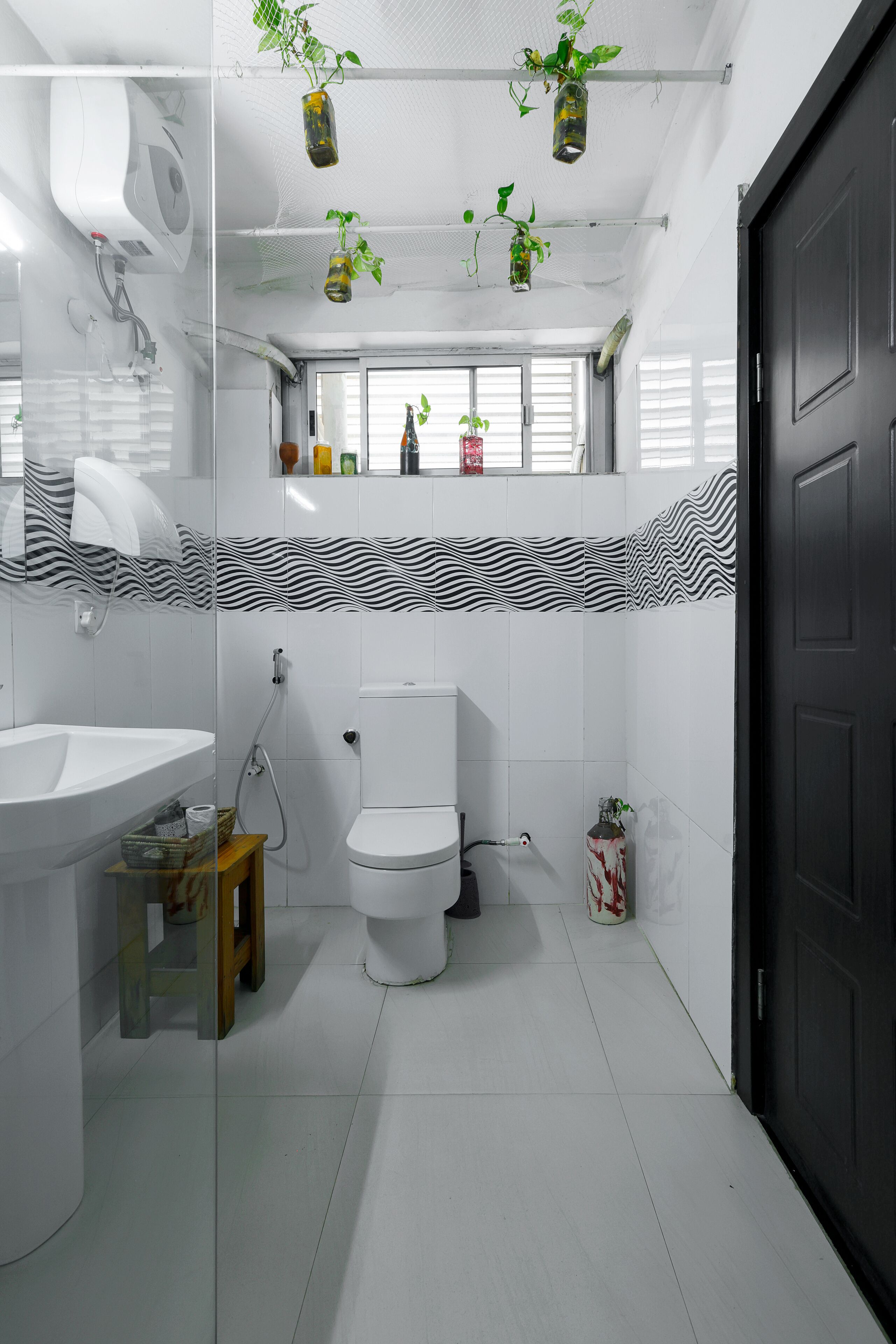 A modern bathroom with a white sink, toilet, and shower. There are hanging plants and a mirror above the sink.