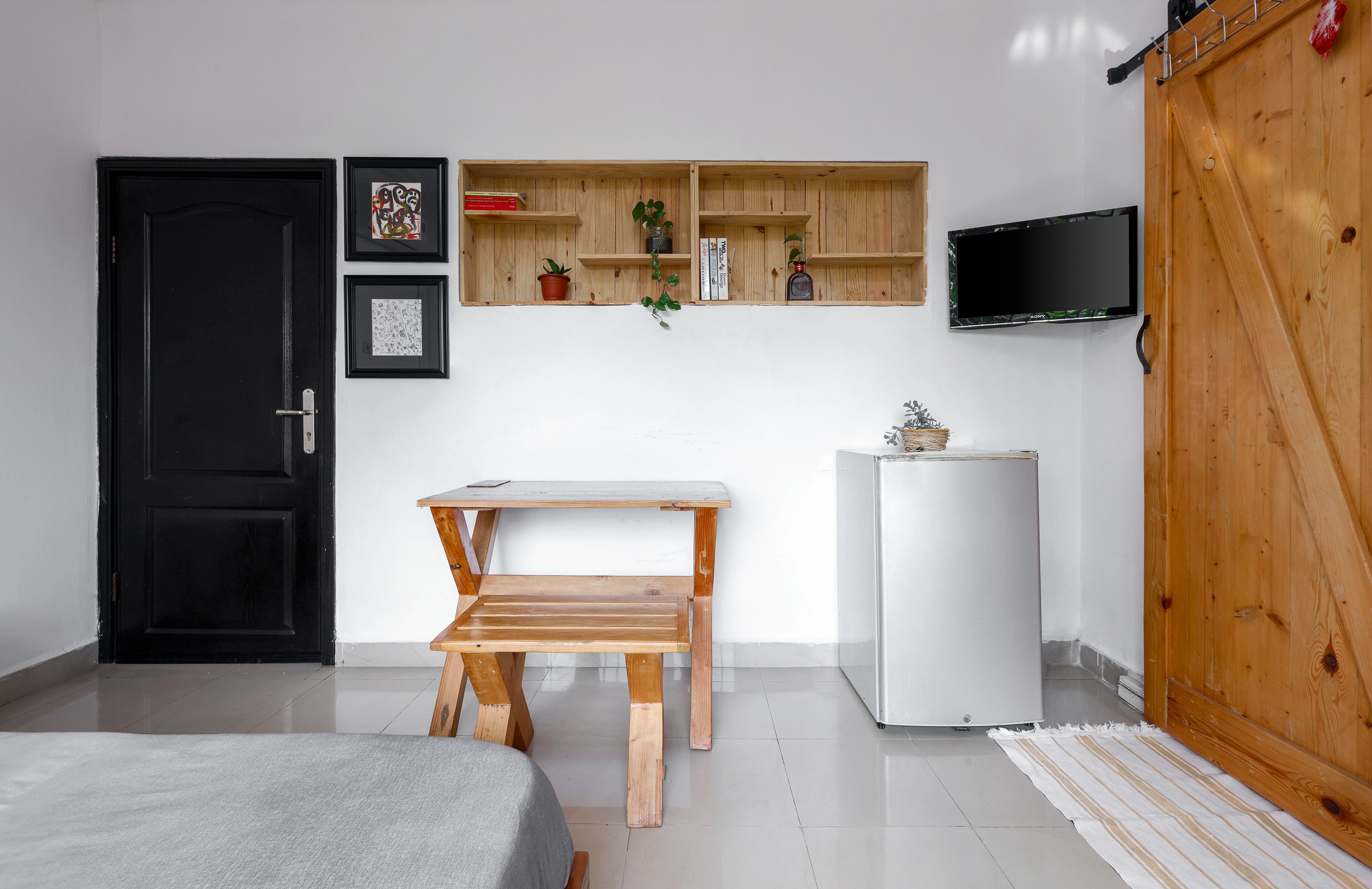 Una habitación con cama, una mesa de madera con un banco, un refrigerador blanco, estantes de madera con libros y plantas, una puerta negra y un televisor montado en la pared.