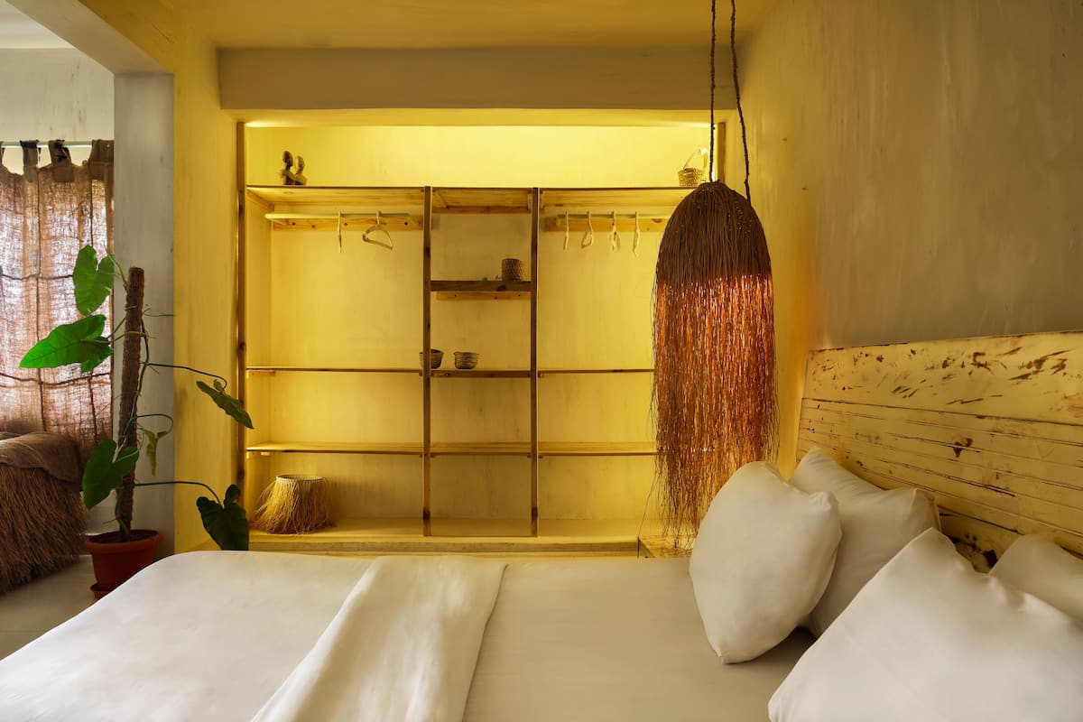 A bedroom with a bed, pillows, a hanging plant, a wooden headboard, and a shelf with items.