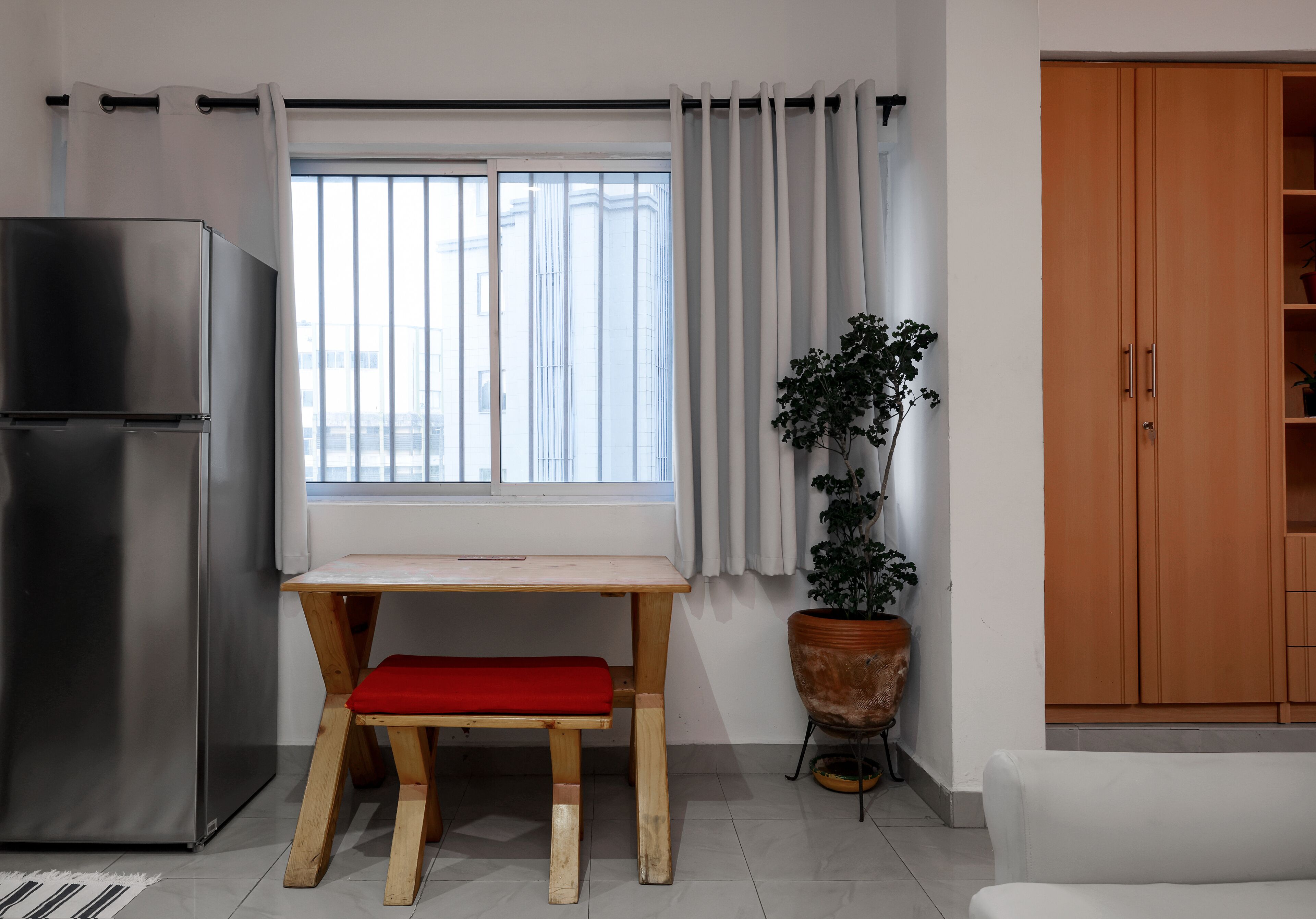 Una cocina compacta con un refrigerador de acero inoxidable, una mesa de madera con un banco rojo, una planta en maceta y un armario.