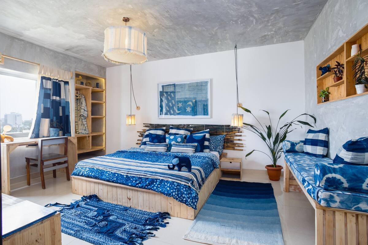 A bedroom with a bed, bedside tables, a desk, and a chair. There is a window with curtains, a shelf with decorative items, and a potted plant.