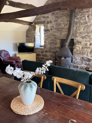 Un interior rústico con pared de piedra, una mesa de madera con un camino tejido, un jarrón con flores y un sofá verde.