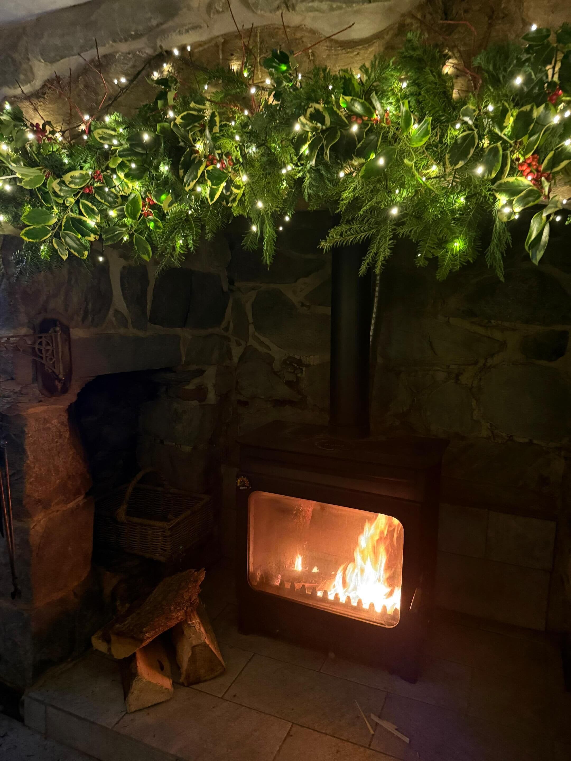 Een stenen open haard met daarin een vuur, omringd door een stenen muur en een decoratieve guirlande met lichtjes.