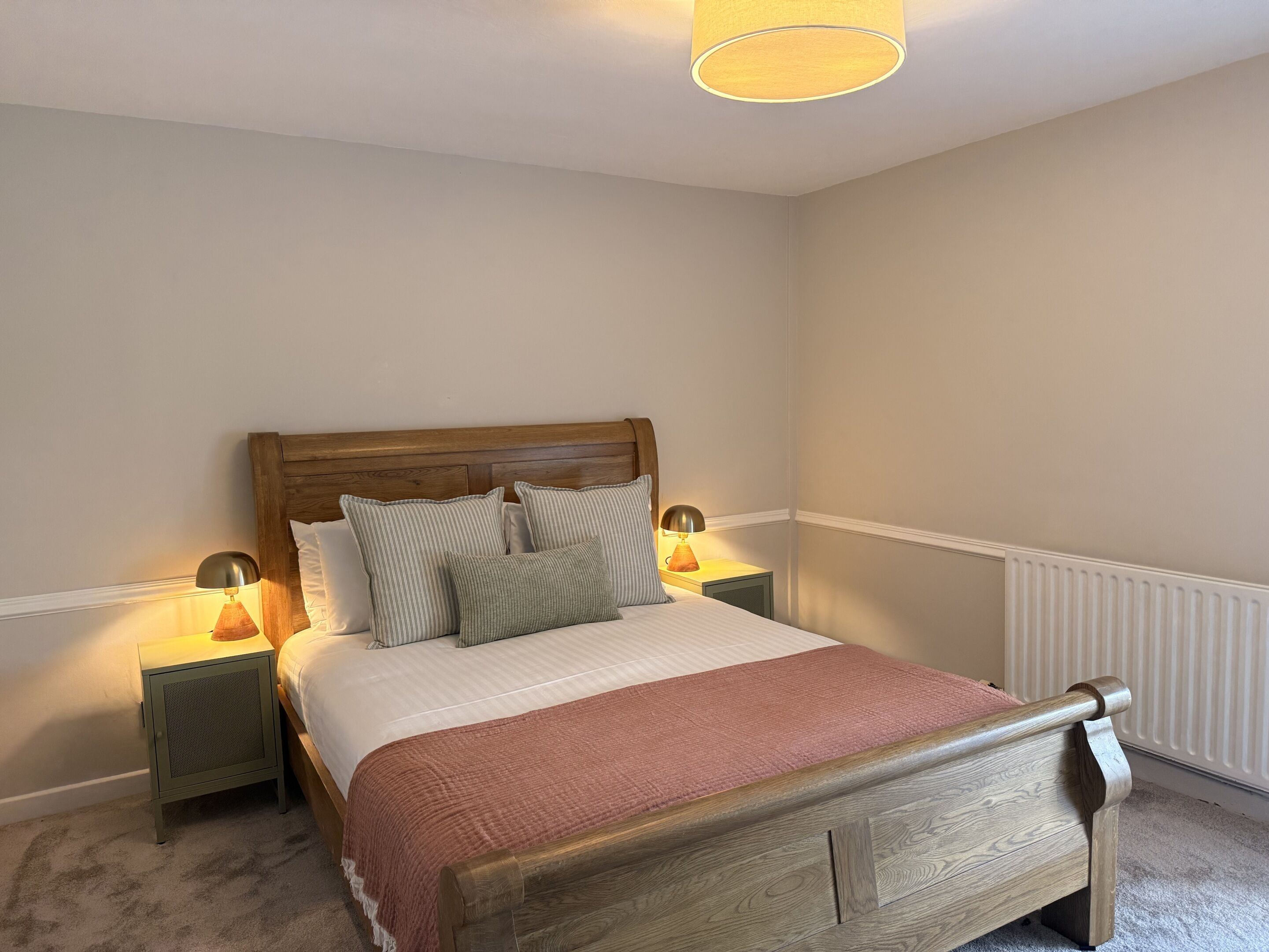 Une chambre à coucher avec un lit en bois, deux tables de chevet avec des lampes, un radiateur et un mur uni.