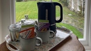 A tray with a mug, a kettle, a jar, and a box of tea on a table by a window with a view of a garden and buildings. - Maes Y Neuadd Country House (Talsarnau)