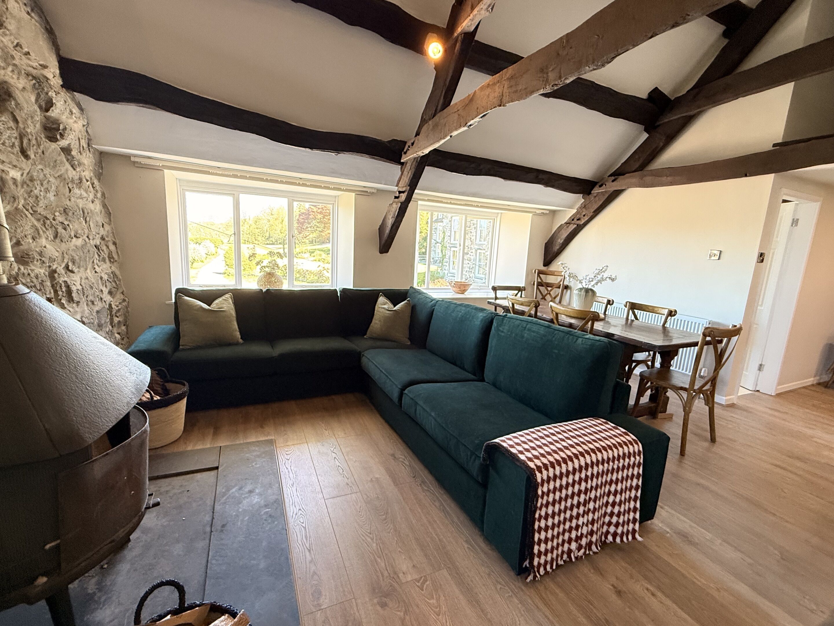 Een kamer met een grote, groene hoekbank, een eettafel met stoelen, een stenen muur en een houten plafond.