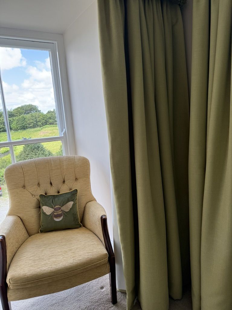 Ein Zimmer mit großem Fenster, ein beiger Sessel mit Bienenkissen und grünen Vorhängen.