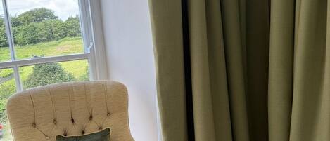 A room with a large window, a beige armchair with a bee pillow, and green curtains.