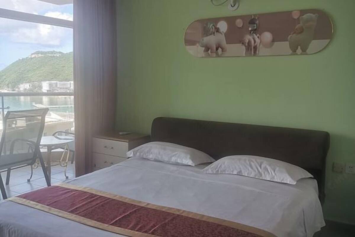 Une chambre à coucher comprenant un lit, une commode, une chaise et donnant sur un plan d’eau et une colline.