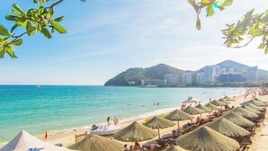 Een strand met rieten parasols, een heldere blauwe zee en een stadsgezicht op de achtergrond.