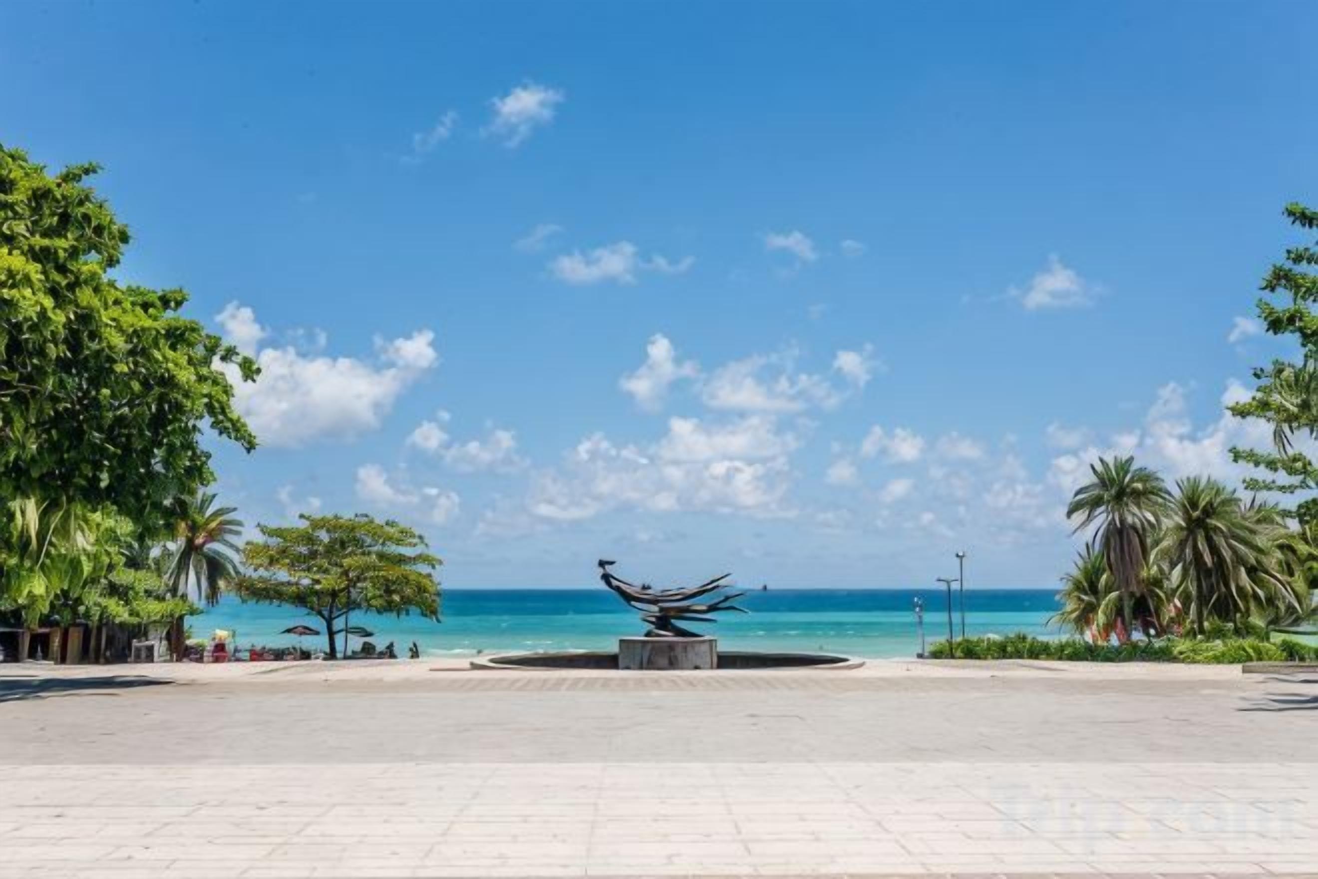 A plaza with a central sculpture, a clear sky, and a view of the ocean.