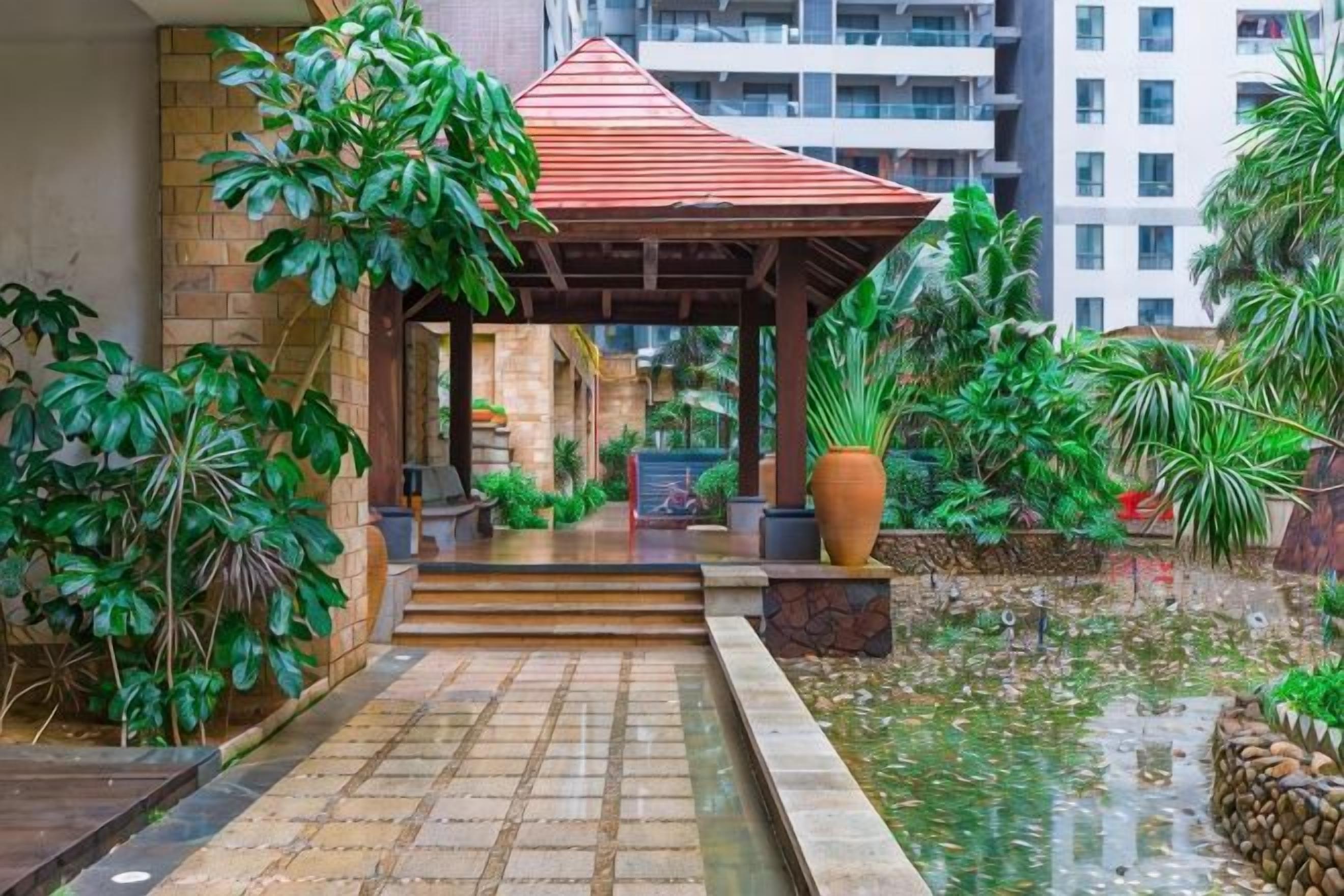 A covered outdoor area with steps leading to a building entrance, surrounded by greenery and a water feature.
