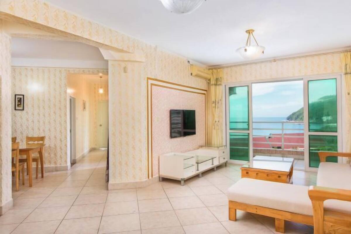 Un salon spacieux doté d’une grande porte-fenêtre coulissante donnant sur un balcon, d’un téléviseur à écran plat fixé au mur et d’un banc en bois.