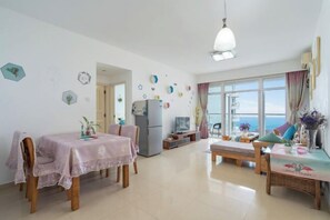 A spacious living area with a dining table, a sofa, and a TV. There is a large window offering a view of the sea. - Sanya Hai Hua Apartment (Sanya)