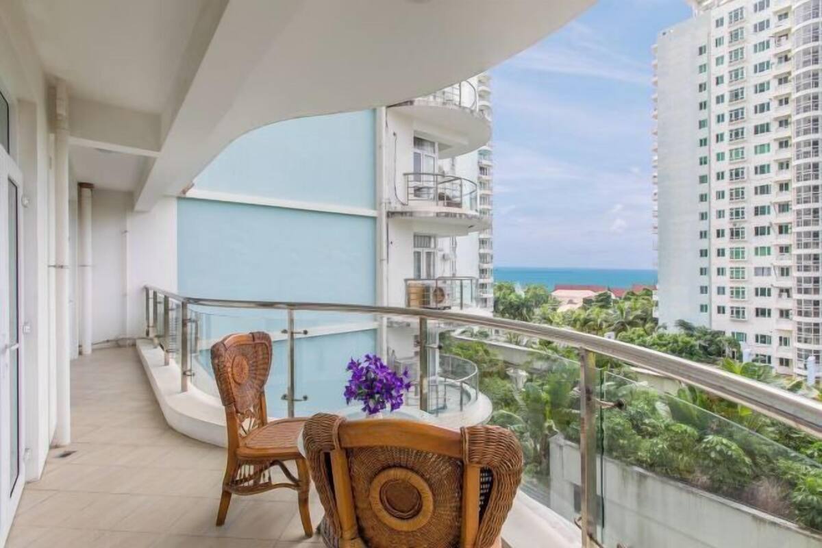 Un balcon avec des fauteuils en osier et une table en verre, offrant une vue sur le paysage urbain et l’océan.