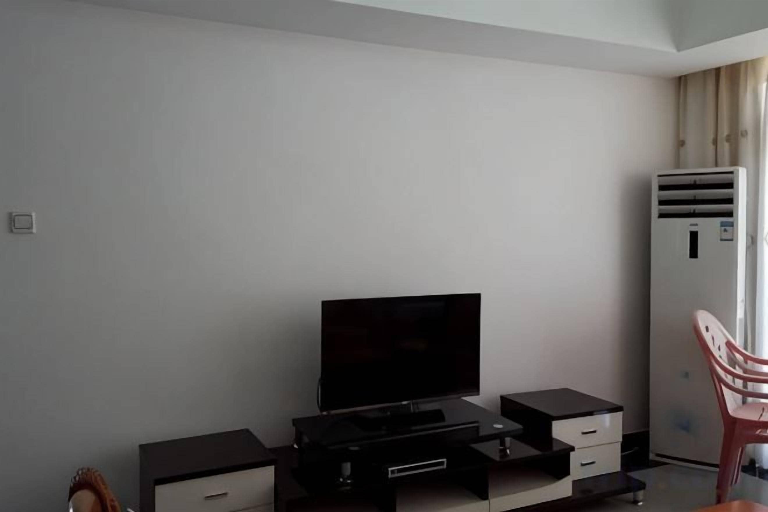 Habitación con televisión de pantalla plana, un escritorio pequeño con cajones, una silla y un refrigerador.