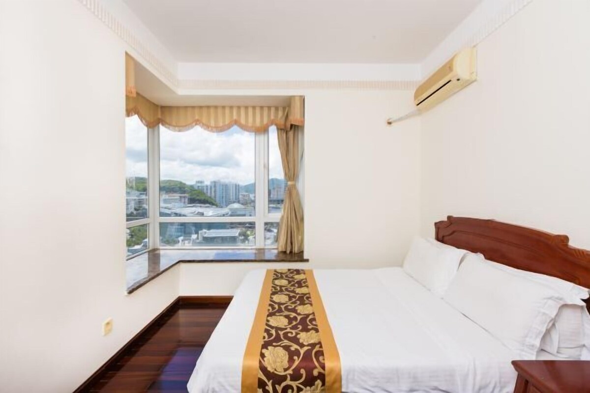A hotel room with a large bed, a window with a view of the city, and an air conditioning unit on the wall.