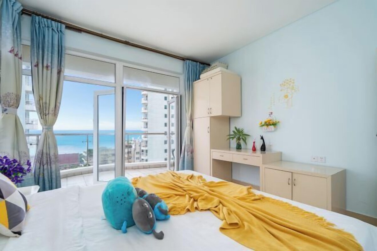 A hotel room with a bed, a yellow blanket, a blue and gray plush toy, a small cabinet, and a view of the sea through a sliding door.