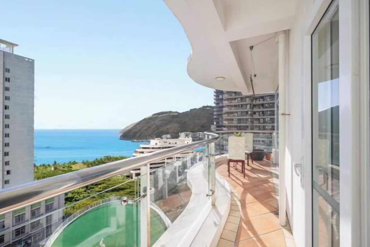 Un balcon doté d’une balustrade en verre, offrant une vue sur la côte, une piscine et un espace repas avec des chaises et une table.
