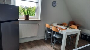 A kitchen with a refrigerator, a dining table with chairs, a clock on the wall, and a window with blinds. - Neue und Ruhig Gelegene Ferienwohnung am Wittensee und in den Hüttener Bergen (Klein Wittensee)