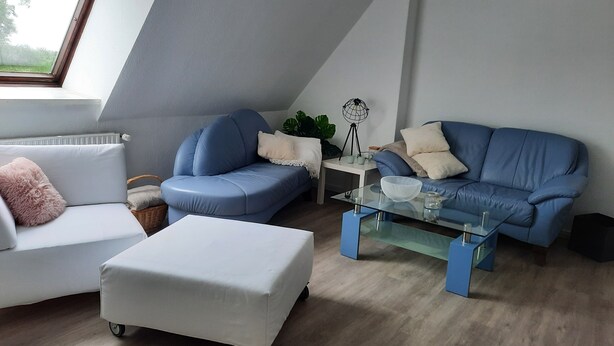 A modern living room with a blue sofa, a white armchair, and a glass coffee table. - Neue und Ruhig Gelegene Ferienwohnung am Wittensee und in den Hüttener Bergen (Klein Wittensee)