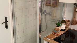 A bathroom with a glass shower enclosure, a wooden shelf with toiletries, and a black countertop with a stove. - Studette Meublée - Boulogne-billancourt Face au Stade de Roland-garros (Boulogne-Billancourt)
